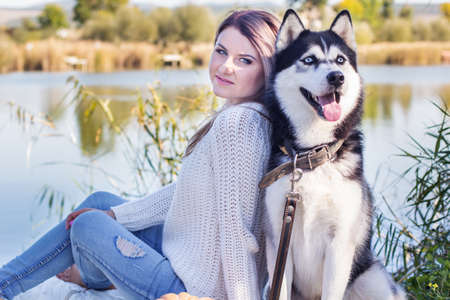 Young girl and her blueyed dog husky is lying outdoors in a park near lake in autumn time