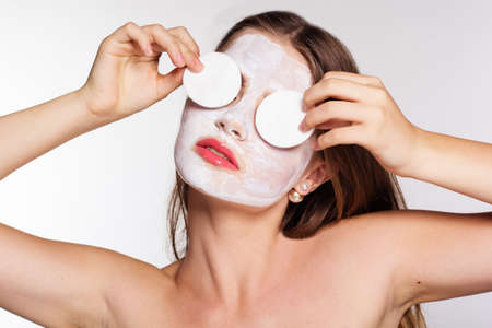 Beautiful smiling women with white clay facial mask on her face is holding cotton pads on hands near eyesの写真素材