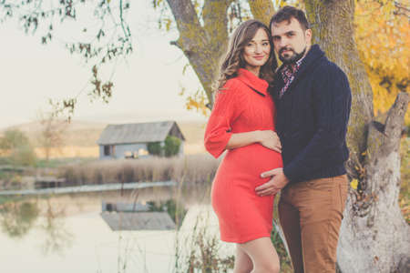 Happy future parents man and pregnant woman are wearing warm clothes spending time in autumn park near big lakeの写真素材