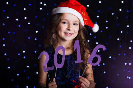 Happy child girl is wearing santa hat holding numbers 2016 in hands in a studio over background...
