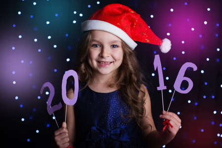 Happy child girl is wearing red santa hat holding numbers 2016 in hands in a studio over background scene with blue lights for a holiday concept.の写真素材