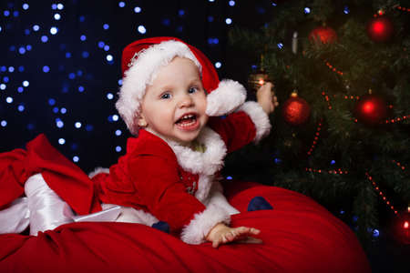 Happy little baby is sitting near fir-tree and holding christmas ball. New Year's holidays. Baby in a red Christmas costume with giftの写真素材