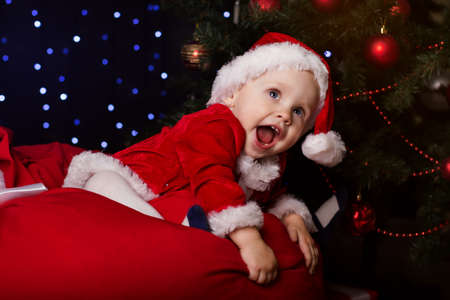 Happy little baby is sitting near fir-tree. New Year's holidays concept. Baby in a red Christmas costume with giftの写真素材