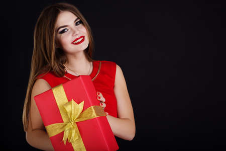 Pretty girl is wearing red dress holding gift box. Smiling beautiful woman portrait isolated on black background.の写真素材