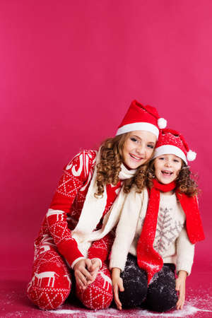 Pretty two sisters girls are blowing fake snow wearing warm winter clothes isolated on red backgroundの写真素材