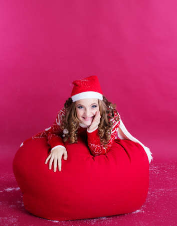 Pretty santa girl is lying on big bag with gifts on her back isolated on red backgroundの写真素材