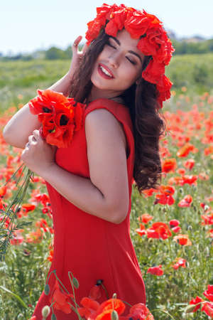 Beautiful smiling girl in poppy field is wearing red fashion dress and wreath of poppiesの写真素材