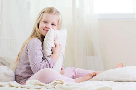 Happy blonde child girl is holding white pillow in the bedの写真素材