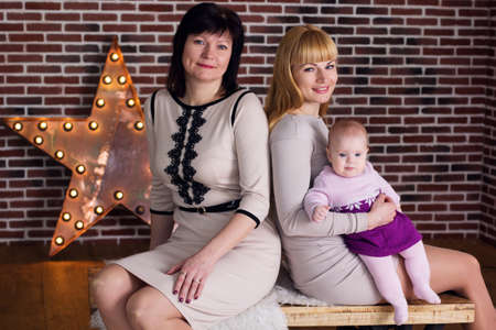 Three steps of family generation: grandmother, mother and granddaughter are sitting over brick wall...