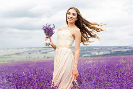 Beautiful smiling woman is wearing nice wedding dress playing with bouquet at field of purple lavender flowersの写真素材