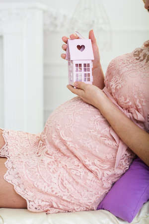 Pretty pregnant woman is holding small pink decorative house in hands