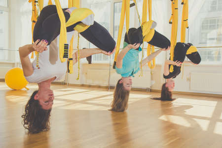 Group of young women doing aerial yoga exercise or antigravity yoga in fitness studio. Fitness, stretch, exercise and healthy lifestyle people. Girls using yellow hammock.の写真素材