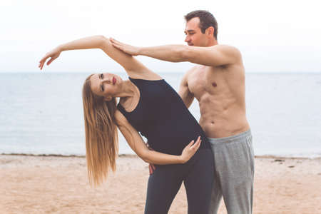 Young sportive couple husband and pregnant wife are doing stretching yoga on the beach, summer timeの写真素材