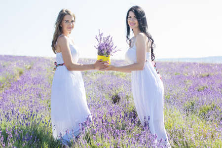 Two young sisters are holding basket in hands and walking on the field of the purple lavender flowers on sunny summer dayの写真素材