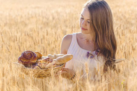 Beautiful teen girl is sitting in a field of rye with basket of fresh bread and sweet bunsの写真素材