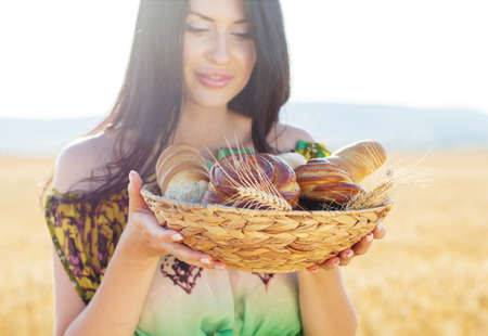 Brunette girl in a field of rye with wicker basket of bread and sweet buns and rollsの写真素材