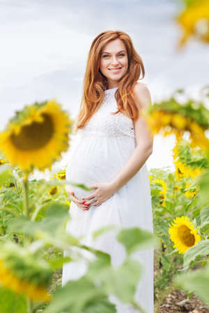 Happy smiling pregnant woman is wearing white fashion dress in field with yellow sunflowersの写真素材