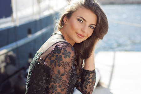 Portrait of pretty girl is wearing fashion dress with luxury makeup sitting near sea with yachtsの写真素材