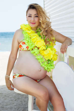 Pregnant woman in sunny summer day is resting on the beach with yellow and green hawaiian flowers on her neckの写真素材