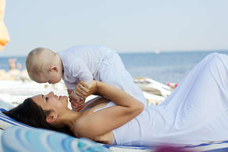 Beautiful smiling mother is wearing white dress with little son are enjoying nice sunny day sitting on sunbed over sea viewの写真素材