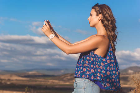 Pretty teen girl is wearing casual clothes taking picture with her phone of mountains and sky view