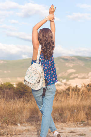 Back view petty traveler teen girl is wearing casual ragged jeans standing with fashion backpack over mountains and sky backgroundの写真素材