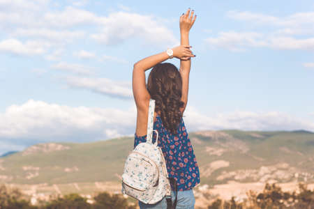 Back view petty traveler teen girl is wearing casual ragged jeans standing with fashion backpack over mountains and sky backgroundの写真素材