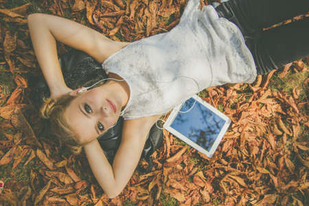 Pretty teenager girl is lying down in autumn park with yellow leaves with digital tablet and...