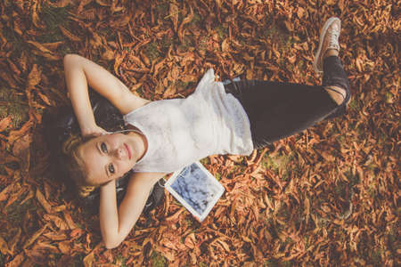 Pretty teenager girl is lying down in autumn park with yellow leaves with digital tablet and...