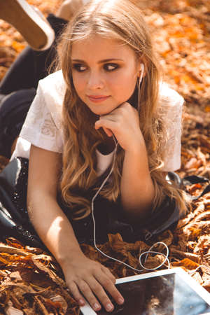 Pretty teenager girl with long hair is lying down in autumn park with yellow leaves with digital...