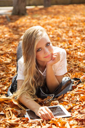 Pretty teenager girl with long hair is lying down in autumn park with yellow leaves with digital tabletの写真素材