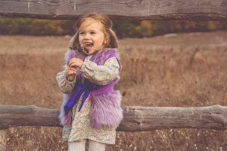 Little child girl is wearing fashion clothes in boho style near wooden fence in autumn fieldの写真素材