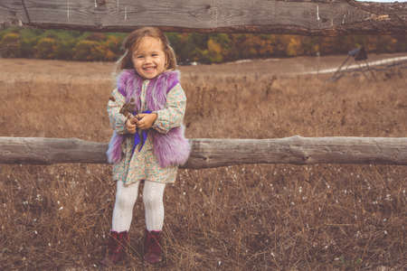 Little child girl is wearing fashion clothes in boho style near wooden fence in autumn fieldの写真素材