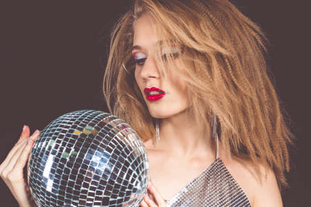 Portrait of attractive blonde woman with fashion makeup is wearing glamour shiny clothes with disco ball over black backgroundの写真素材