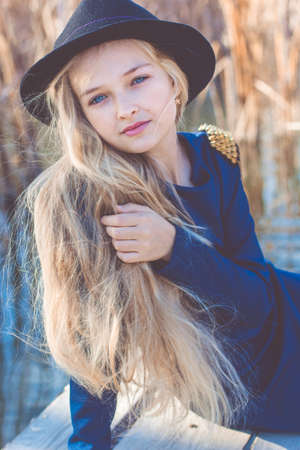 Little cute girl with long blonde hair outdoors is sitting near lake and wearing fashion dress and hatの写真素材