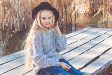 Little cute girl with long blonde hair outdoors is sitting near lake and wearing warm sweater and hatの写真素材