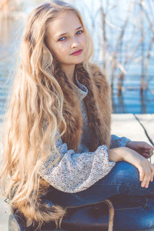 Little cute smiling girl with long blonde hair outdoors is sitting near lake and wearing warm sweater and furry jacketの写真素材