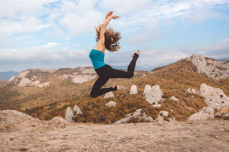 Young pretty flexible woman doing jumping in beautiful mountains landscape view on the top of the hillの写真素材