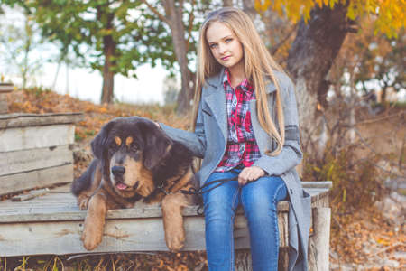 Pretty teen girl with blonde long hair is wearing warm winter clothes sitting with puppy of brown tibetan mastiff in autumn park near lakeの写真素材