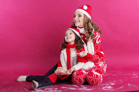 Pretty happy two sisters girls are playing and having fun with fake snow wearing warm red winter clothes isolated on red background, christmas conceptの写真素材