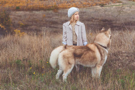 Little pretty child girl is wearing fashion warm light clothes walking around with her hairy husky dog on the nature, autumn seasonの写真素材