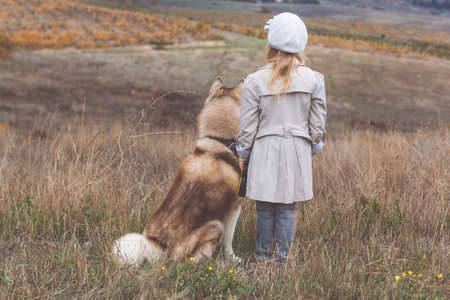 Back view of child girl is wearing fashion warm light clothes with her hairy husky dog on the nature, autumn seasonの写真素材