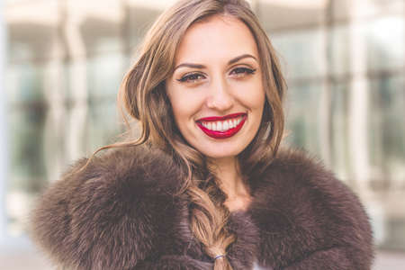 Portrait of beautiful gorgeous smiling woman is wearing luxury dark fur coat posing in near office buildingの写真素材