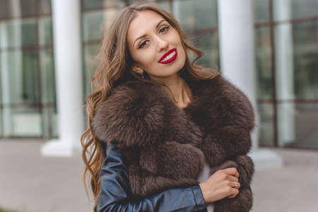 Portrait of beautiful gorgeous smiling woman is wearing luxury dark fur coat posing in near office buildingの写真素材