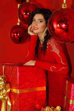Pretty happy brunette woman is wearing red dress in holiday room with huge Christmas gift box with golden ribbon, happy new year conceptの写真素材