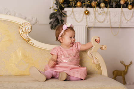 Cute baby girl is wearing pink dress sitting on the sofa near beautiful fireplace with xmas decorationsの写真素材