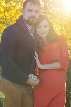 Happy future parents wife and husband are wearing warm clothes spending time in autumn park in sunset lightsの写真素材