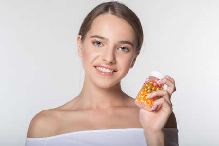 Portrait of young smiling girl with perfect skin and nude makeup isolated on white in studio is holding jar of colorful vitamins in handsの写真素材