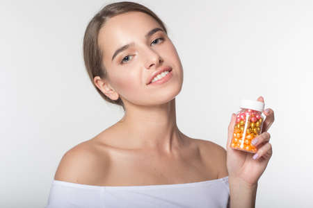 Portrait of young caucasian girl with perfect skin and nude makeup isolated on white in studio is holding jar of colorful vitamins in handsの写真素材