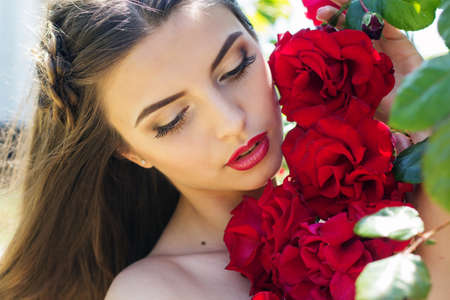 Beautiful young smiling woman with long curly hair posing near red roses in a gardenの写真素材